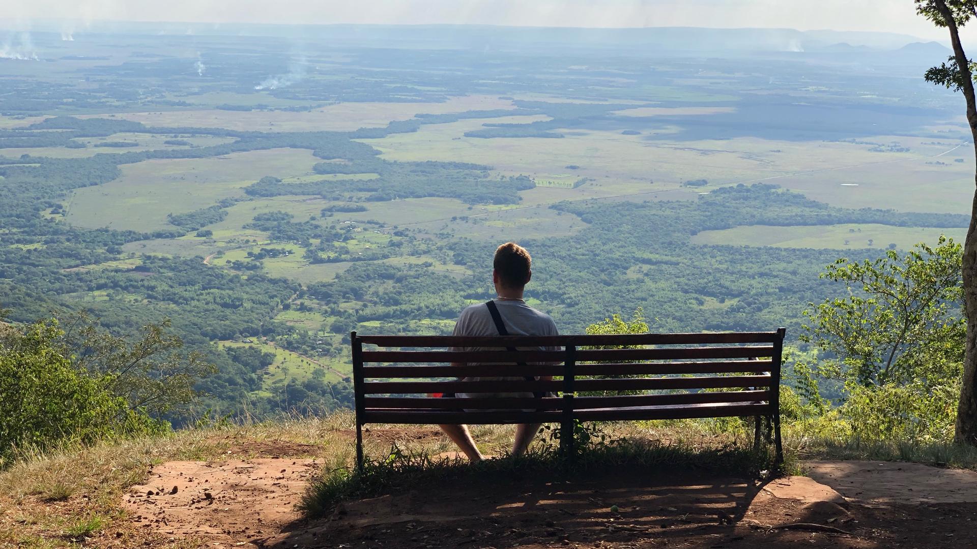 Ein junger Mann sitzt auf einer Bank und schaut über ein großes grünes Tal.