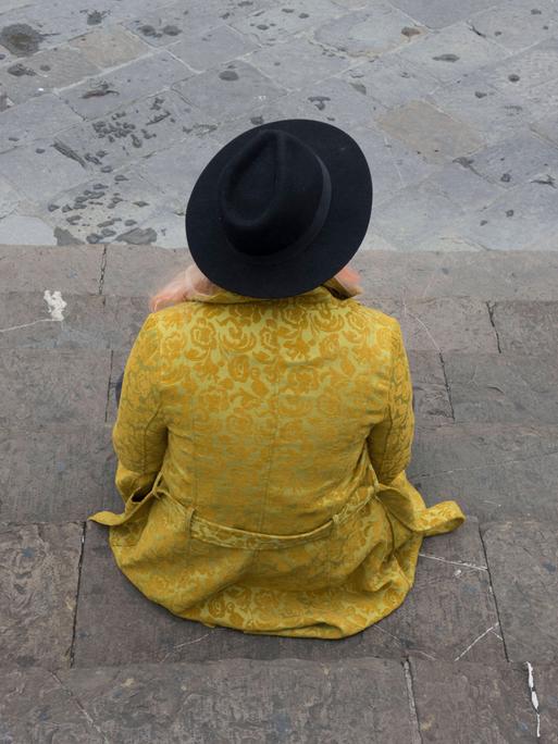 Ein junges Mädchen trägt einen gelben Mantel mit schwarzen Hut und sitzt alleine auf einer Stufe
