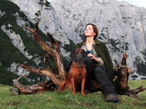 Im Filmstill von "Die Wand" sitzt Martina Gedeck mit ihrem Hund an der Seite auf einem Baumstumpf und schaut ins Gebirge.