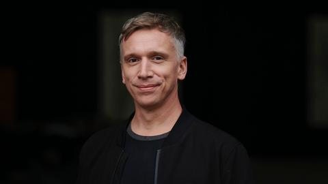 Ein Mann mit kurzen braun-grauen Haaren trägt eine schwarze dünne Jacke und lächelt in die Kamera. Der Hintergrund ist schwarz.