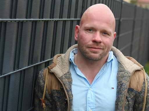 Der Intensivpfleger Ricardo Lange trägt ein hellblaues Hemd und darüber eine offene, braune Jacke und lächelt leicht in die Kamera. Der Intensivpfleger Ricardo Lange trägt ein hellblaues Hemd und darüber eine offene, braune Jacke und lächelt leicht in die Kamera.
