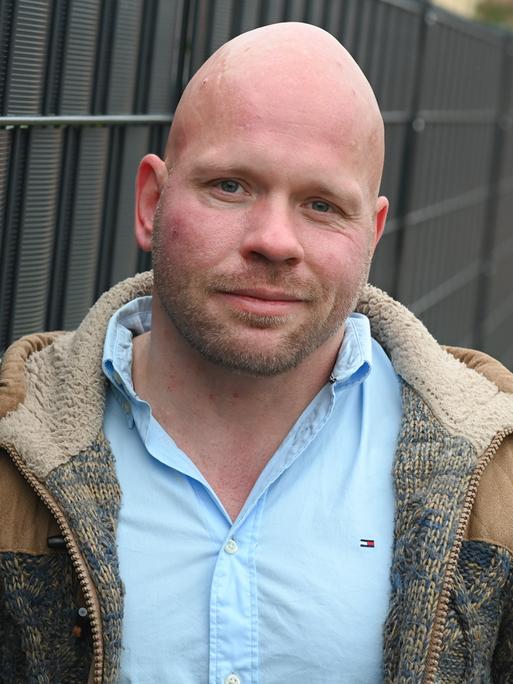Der Intensivpfleger Ricardo Lange trägt ein hellblaues Hemd und darüber eine offene, braune Jacke und lächelt leicht in die Kamera. Der Intensivpfleger Ricardo Lange trägt ein hellblaues Hemd und darüber eine offene, braune Jacke und lächelt leicht in die Kamera.