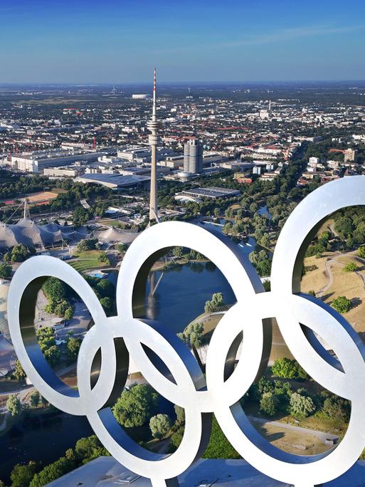 Luftaufnahme des Olympiaparks mit Olympiastadion, Olympia-Schwimmhalle und Olympiahalle, davor die Olympischen Ringe auf einer Dachkonstruktion (Fotomontage) 