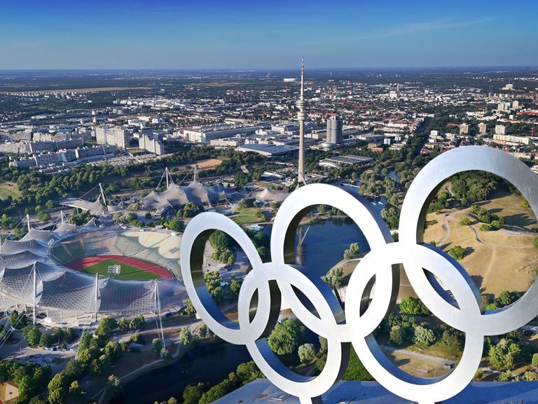 Luftaufnahme des Olympiaparks mit Olympiastadion, Olympia-Schwimmhalle und Olympiahalle, davor die Olympischen Ringe auf einer Dachkonstruktion (Fotomontage) 