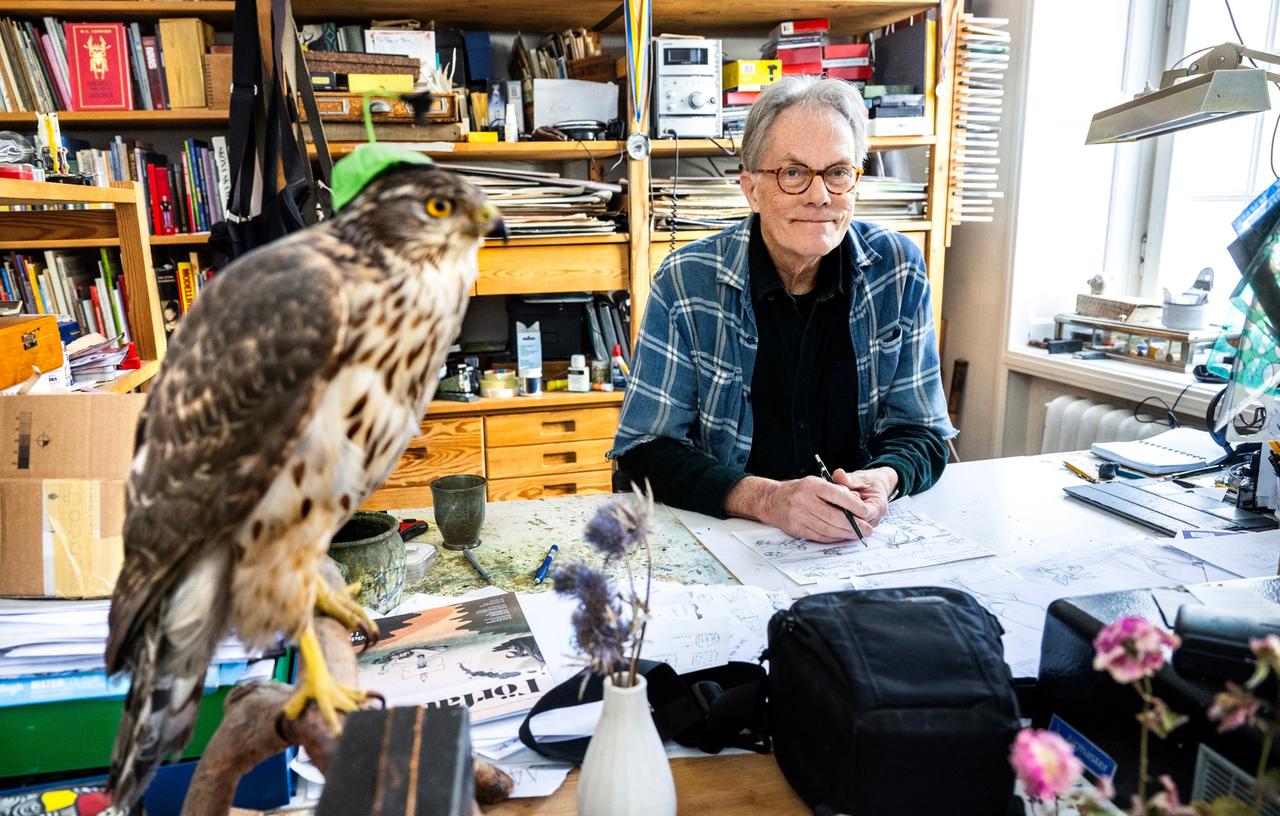 Der Illustrator und Kinderbuchautor Sven Nordqvist sitzt hinter seinem Schreibtisch in seinem Atelier in Stockholm. Hinter ihm steht ein mit Büchern, Zeichenmappen und Nippes gefülltes Regal. Im Vordergrund hockt eine ausgestopfte Eule auf einem Stock.