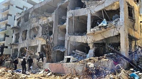 Menschen stehen vor den Trümmern eines Wohnhauses. Die Fassade ist abgesprengt, Wände sind schief.