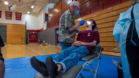 In einer Turnhalle liegt ein junger Mann auf einem provisorischen Zahnarztstuhl und wird untersucht. In einer Turnhalle liegt ein junger Mann auf einem provisorischen Zahnarztstuhl und wird untersucht.