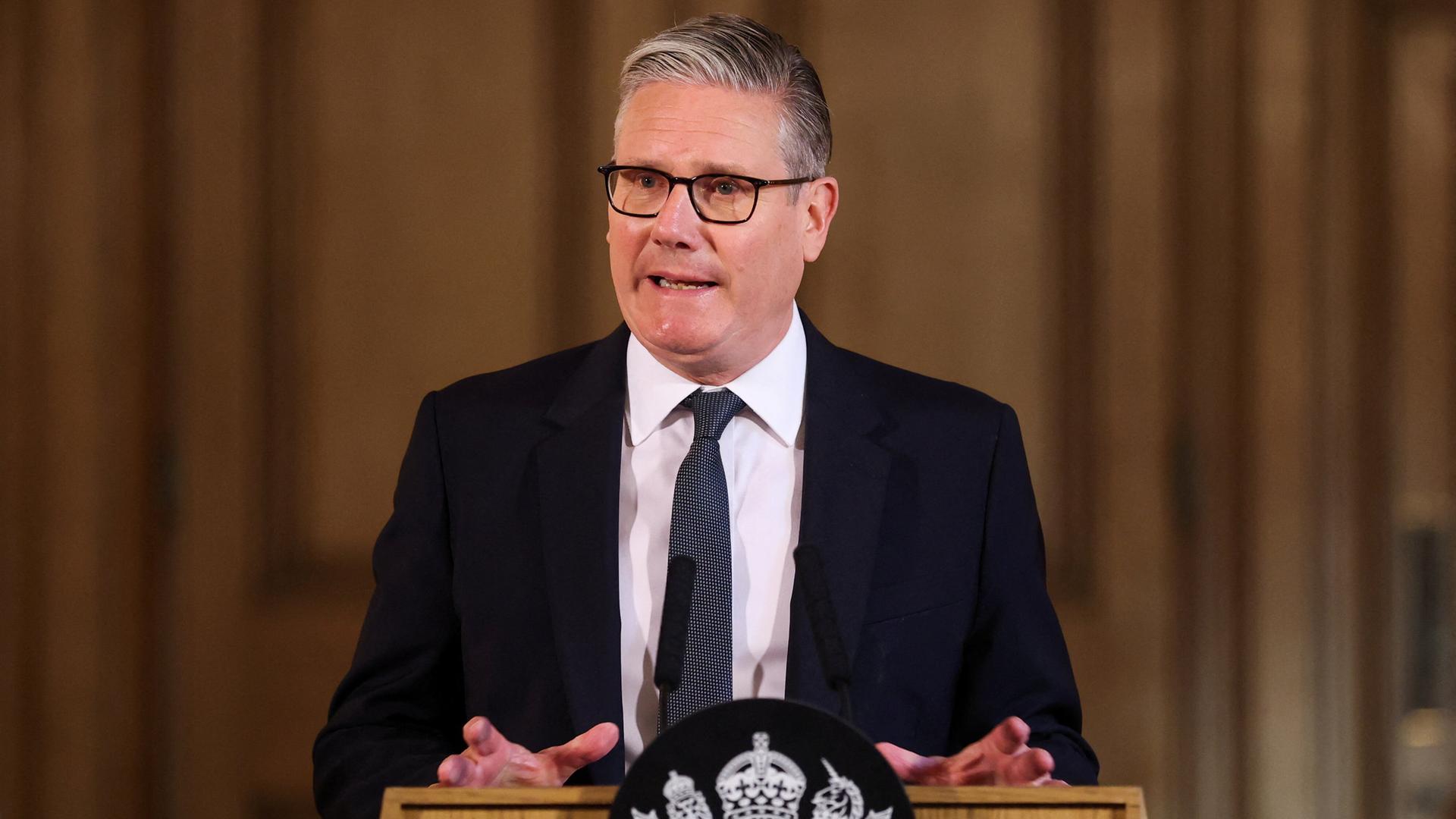 Keir Starmer steht an einem Redner-Pult und spricht. Er trägt einen schwarzen Anzug und eine Krawatte.