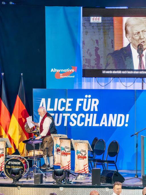 Über der Bühne bei einer Wahlkampfveranstaltung mit Deutschlandflaggen und AfD-Logos ist eine Leinwand mit einem Wahlkampfvideo von Donald Trump zu sehen Über der Bühne bei einer Wahlkampfveranstaltung mit Deutschlandflaggen und AfD-Logos ist eine Leinwand mit einem Wahlkampfvideo von Donald Trump zu sehen