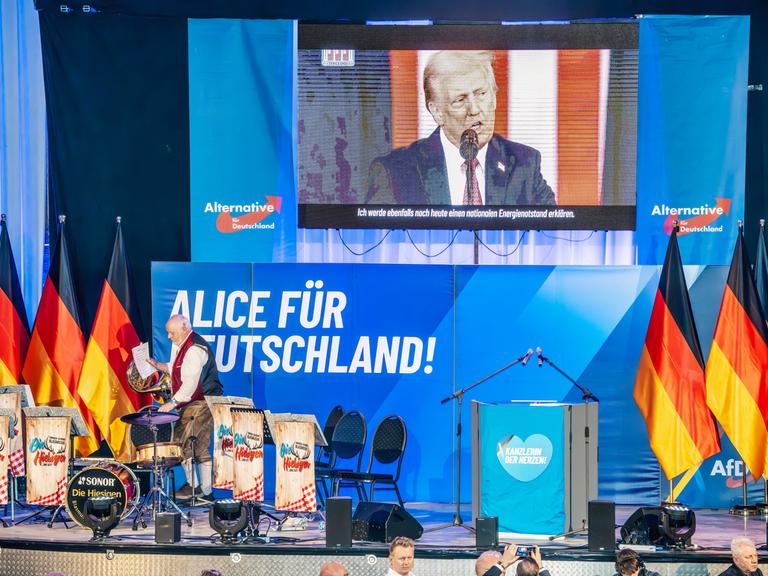 Über der Bühne bei einer Wahlkampfveranstaltung mit Deutschlandflaggen und AfD-Logos ist eine Leinwand mit einem Wahlkampfvideo von Donald Trump zu sehen Über der Bühne bei einer Wahlkampfveranstaltung mit Deutschlandflaggen und AfD-Logos ist eine Leinwand mit einem Wahlkampfvideo von Donald Trump zu sehen