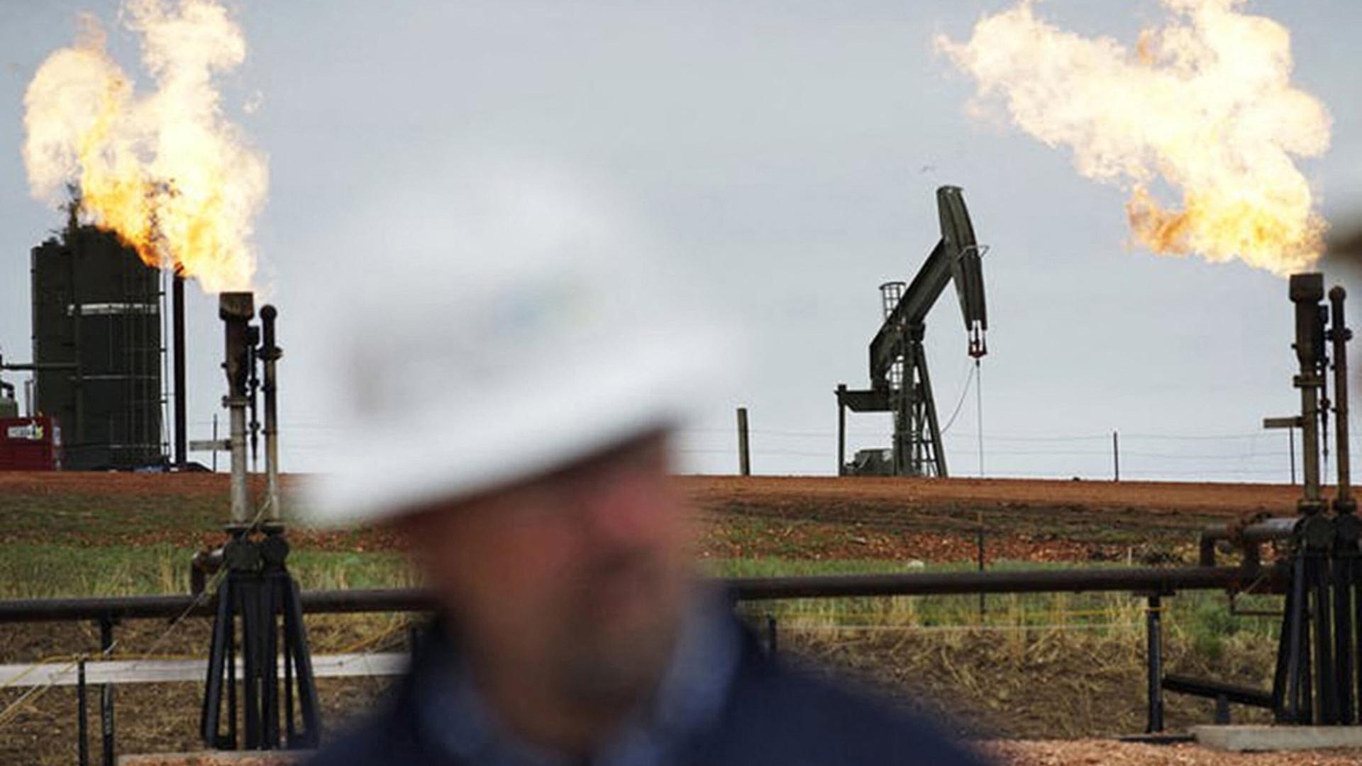 Foto der Erdölförderanlage im US-Bundesstaat North Dakota. Ein Mann im Vordergrund trägt einen weißen Schutzhelm, im Hintergrund sind Förderpumpen und brennende Schornsteine.