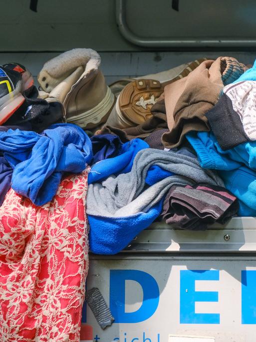 Kleidungsstücke und Schuhe liegen auf einem übervollen Altkleidercontainer.