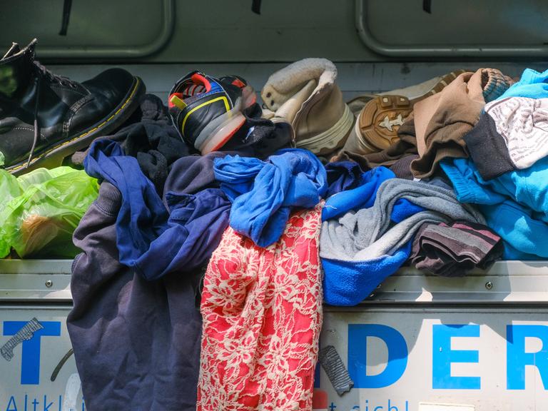 Kleidungsstücke und Schuhe liegen auf einem übervollen Altkleidercontainer.