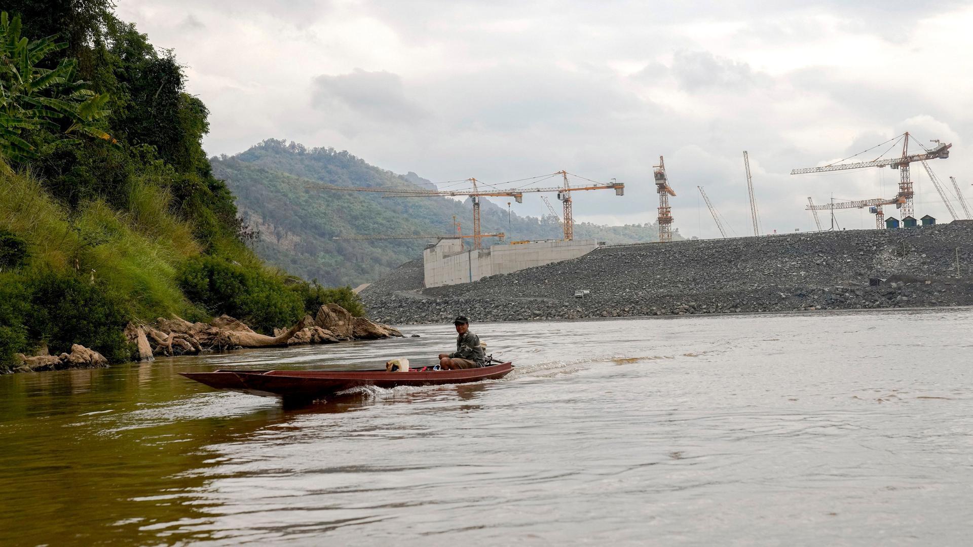 Ein Boot auf dem Mekong bei Luang Prabang in Laos. Im Hintergrund Kräne an der Baustelle für einen Staudamm. Ein Boot auf dem Mekong bei Luang Prabang in Laos. Im Hintergrund Kräne an der Baustelle für einen Staudamm.