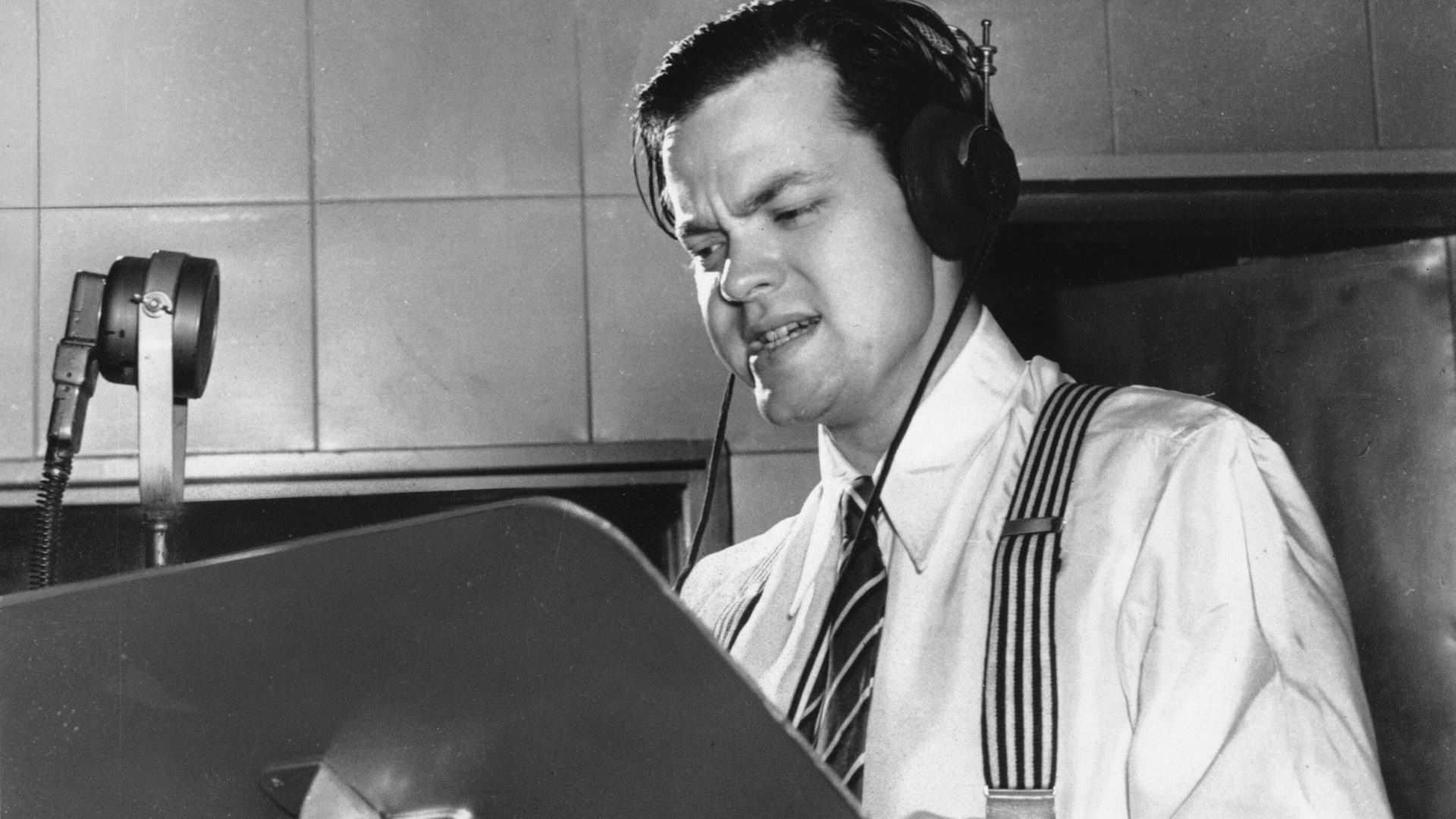 Orson Welles steht in dramatischer Pose an einem Rednerpult in einem Studio und nimmt ein Hörspiel auf.