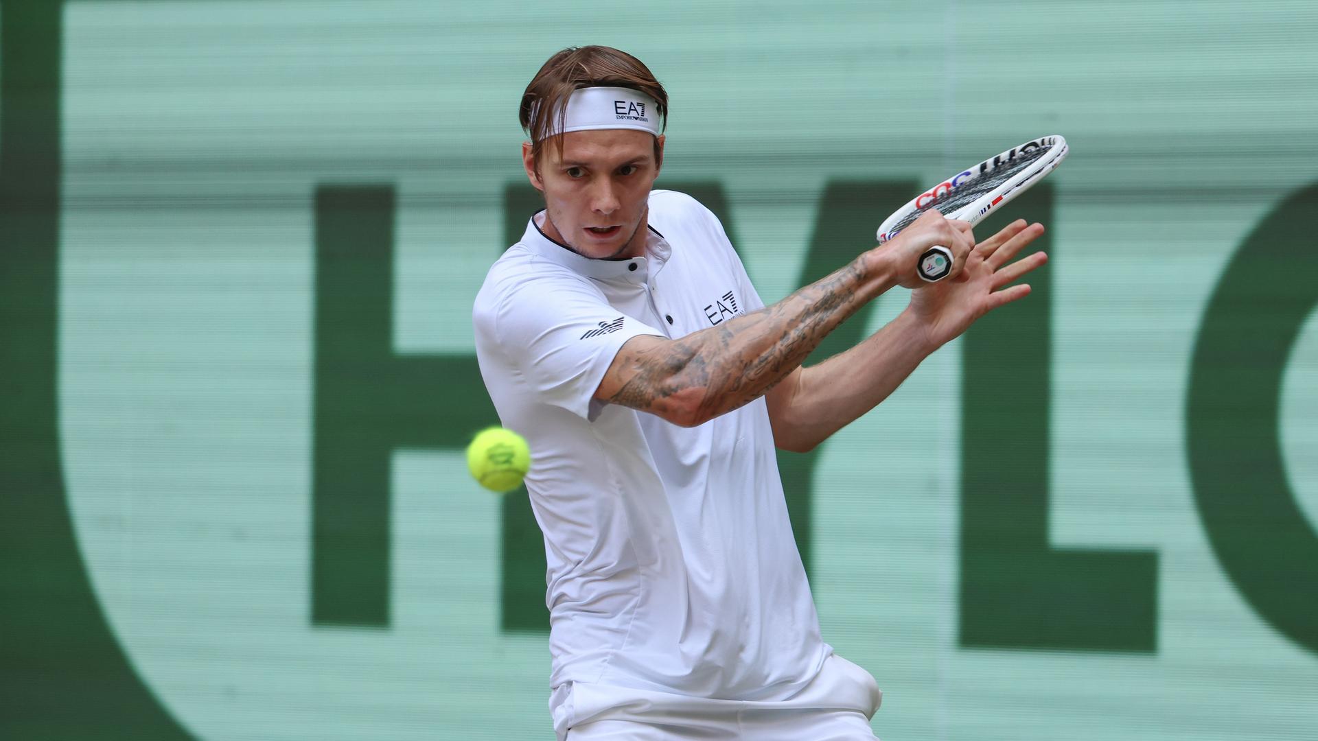 Alexander Bublik spielt eine Rückhand. Er trägt ein weißes Tennisshirt und ein weißes Stirnband. Sein rechter Arm ist tätowiert. Alexander Bublik spielt eine Rückhand. Er trägt ein weißes Tennisshirt und ein weißes Stirnband. Sein rechter Arm ist tätowiert.