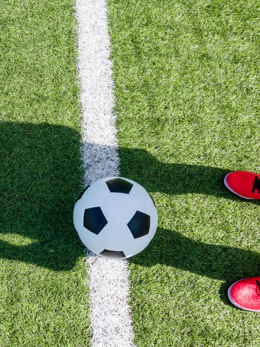 Auf einem Fussballfeld liegt ein Fussball und eine Person mit roten Schuhen wirft einen Schatten auf das Feld und den Ball. Auf einem Fussballfeld liegt ein Fussball und eine Person mit roten Schuhen wirft einen Schatten auf das Feld und den Ball.
