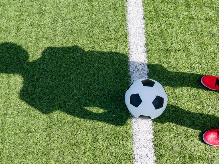 Auf einem Fussballfeld liegt ein Fussball und eine Person mit roten Schuhen wirft einen Schatten auf das Feld und den Ball. Auf einem Fussballfeld liegt ein Fussball und eine Person mit roten Schuhen wirft einen Schatten auf das Feld und den Ball.