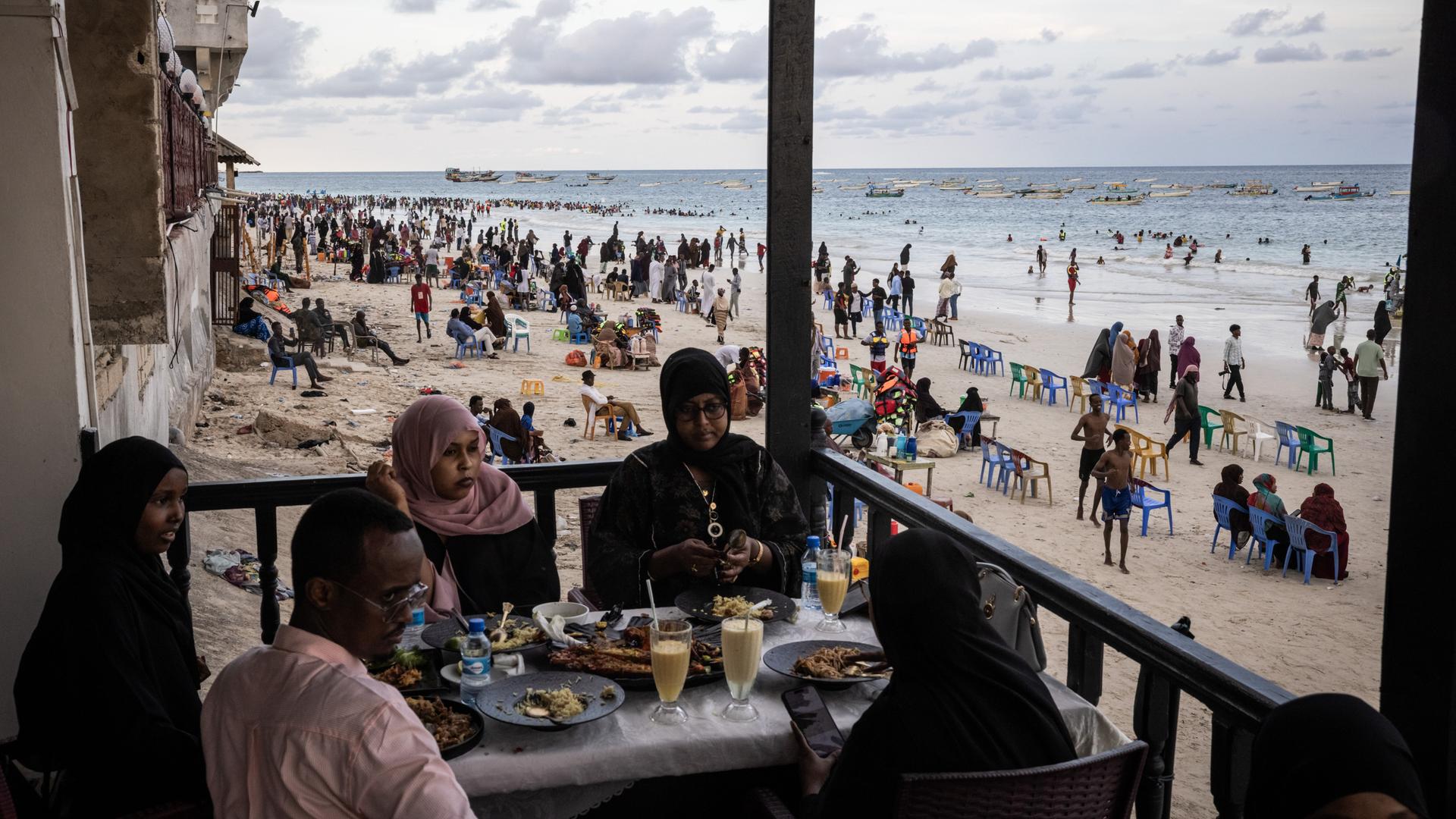 Gäste sitzen in einem Restaurant am Lido Beach von Mogadischu in Somalia, während im Hintergrund am Strand sehr viele Menschen ihre Freizeit verbringen. Gäste sitzen in einem Restaurant am Lido Beach von Mogadischu in Somalia, während im Hintergrund am Strand sehr viele Menschen ihre Freizeit verbringen.