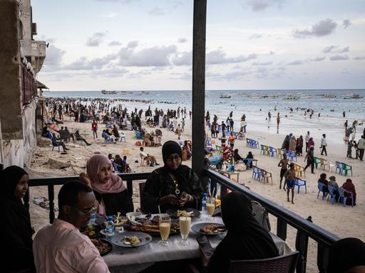 Gäste sitzen in einem Restaurant am Lido Beach von Mogadischu in Somalia, während im Hintergrund am Strand sehr viele Menschen ihre Freizeit verbringen.