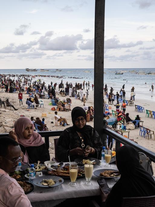 Gäste sitzen in einem Restaurant am Lido Beach von Mogadischu in Somalia, während im Hintergrund am Strand sehr viele Menschen ihre Freizeit verbringen.