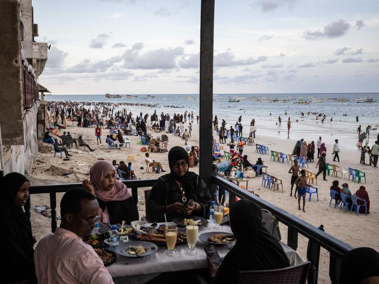 Gäste sitzen in einem Restaurant am Lido Beach von Mogadischu in Somalia, während im Hintergrund am Strand sehr viele Menschen ihre Freizeit verbringen.