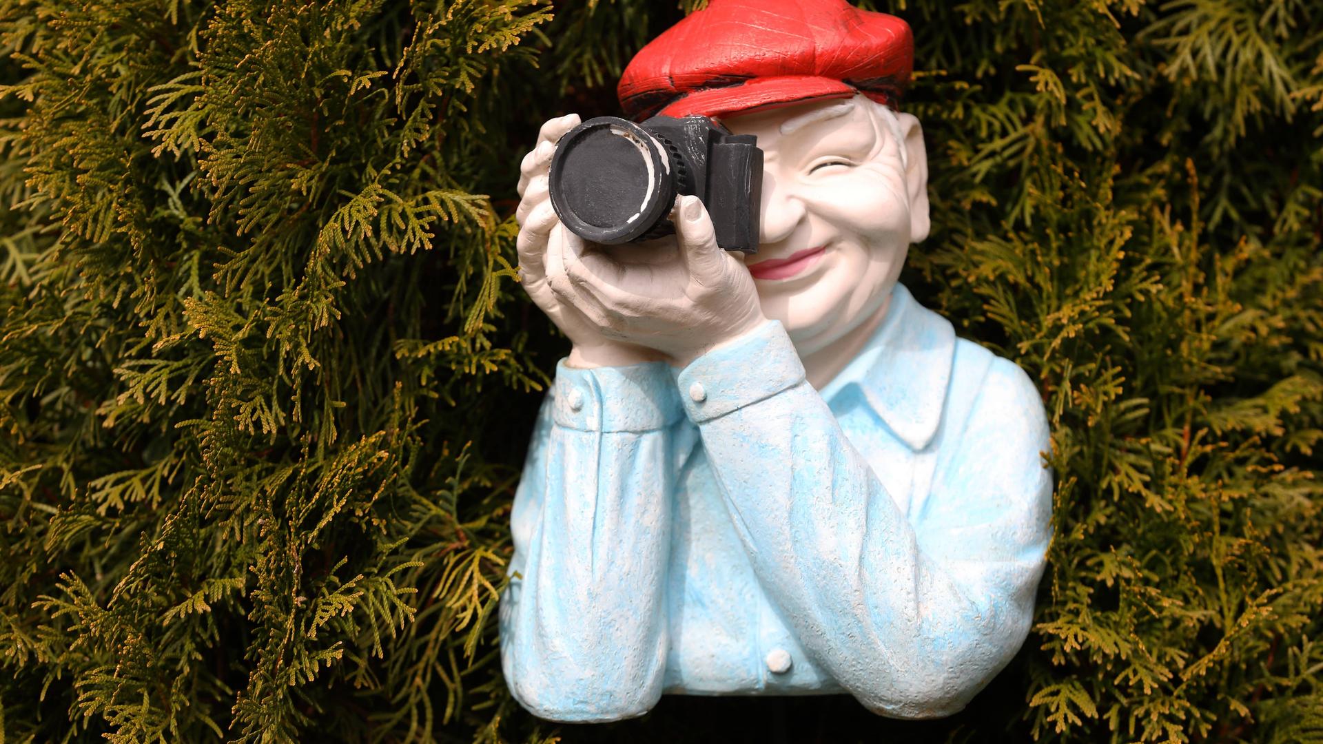Mann in einer Nachbildung aus Kunststoff trägt eine rote Mütze mit einer schwarzen Kamera positioniert vor einer Hecke. Mann in einer Nachbildung aus Kunststoff trägt eine rote Mütze mit einer schwarzen Kamera positioniert vor einer Hecke.