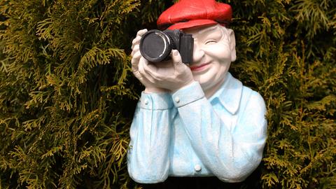 Mann in einer Nachbildung aus Kunststoff trägt eine rote Mütze mit einer schwarzen Kamera positioniert vor einer Hecke.