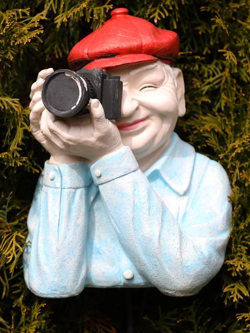 Mann in einer Nachbildung aus Kunststoff trägt eine rote Mütze mit einer schwarzen Kamera positioniert vor einer Hecke.