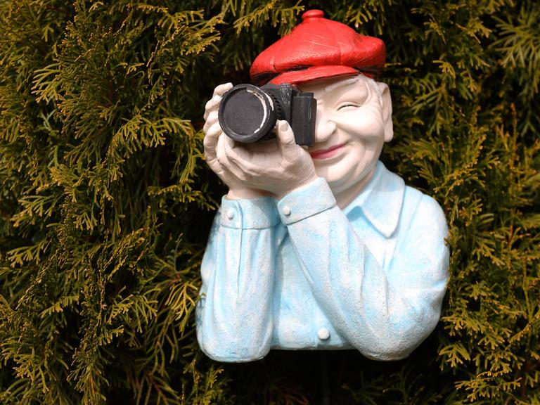 Mann in einer Nachbildung aus Kunststoff trägt eine rote Mütze mit einer schwarzen Kamera positioniert vor einer Hecke.