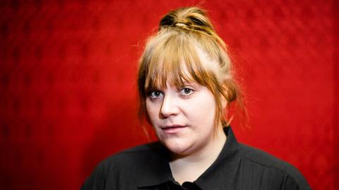 Stefanie Reinsperger steht vor einem roten Hintergrund und blickt in die Kamera. 