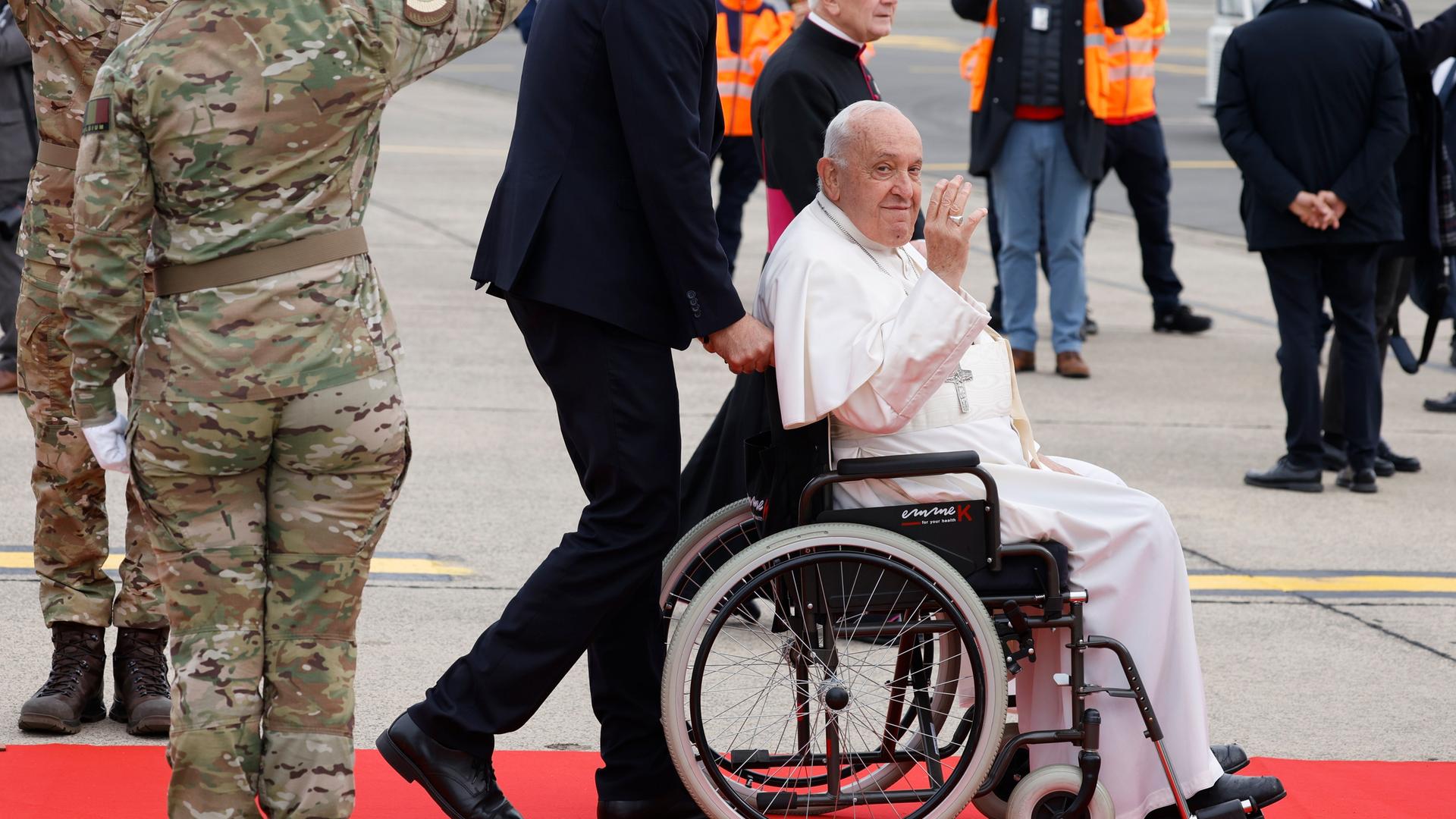 Papst Franziskus auf dem Flughafen in der Nähe von Brüssel kurz vor seinem Abflug nach Rom: Der 87-Jährige winkt aus seinem Rollstuhl, neben ihm stehen eine Soldatin und ein Soldat und salutieren. 