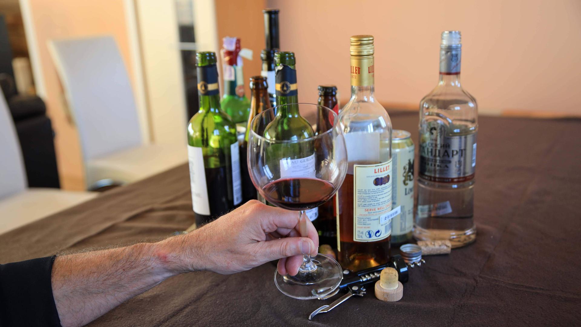 Viele Flaschen mit Alkohol stehen auf einem Tisch. Eine Hand hält ein Glas Rotwein vorne ins Bild.