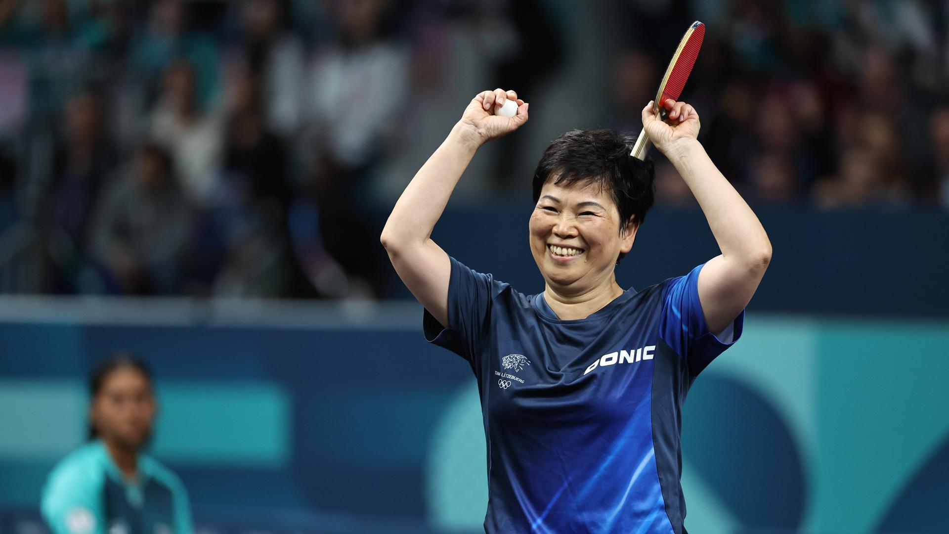 Ein Frau mit kurzen schwarzen Haaren, bekleidet mit einem blau-schwarzem Sportshirt, reckt die Arme hoch und lächelt. Sie hält Tischtennisschläger und  Tischtennisball in der Hand.