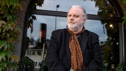 Porträt von Jon Fosse. Er steht vor einem efeuumrankten Fenster, trägt ein schwarzes Jacket und braun gemustertes Halstuch.