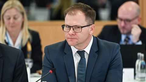 Carsten Schneider (SPD) sitzt an einem Tisch, hinter ihm verschwommen ein Mann und eine Frau.