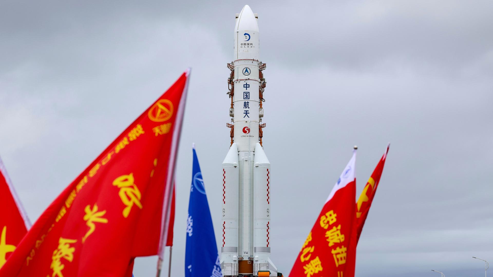 Die Mondsonde Chang'e-7 und ihre Trägerrakete am 19. August 2026 in der südchinesischen Provinz Hainan; im Vordergrund sind rote und blaue Fahnen mit chinesischen Schriftzeichen.