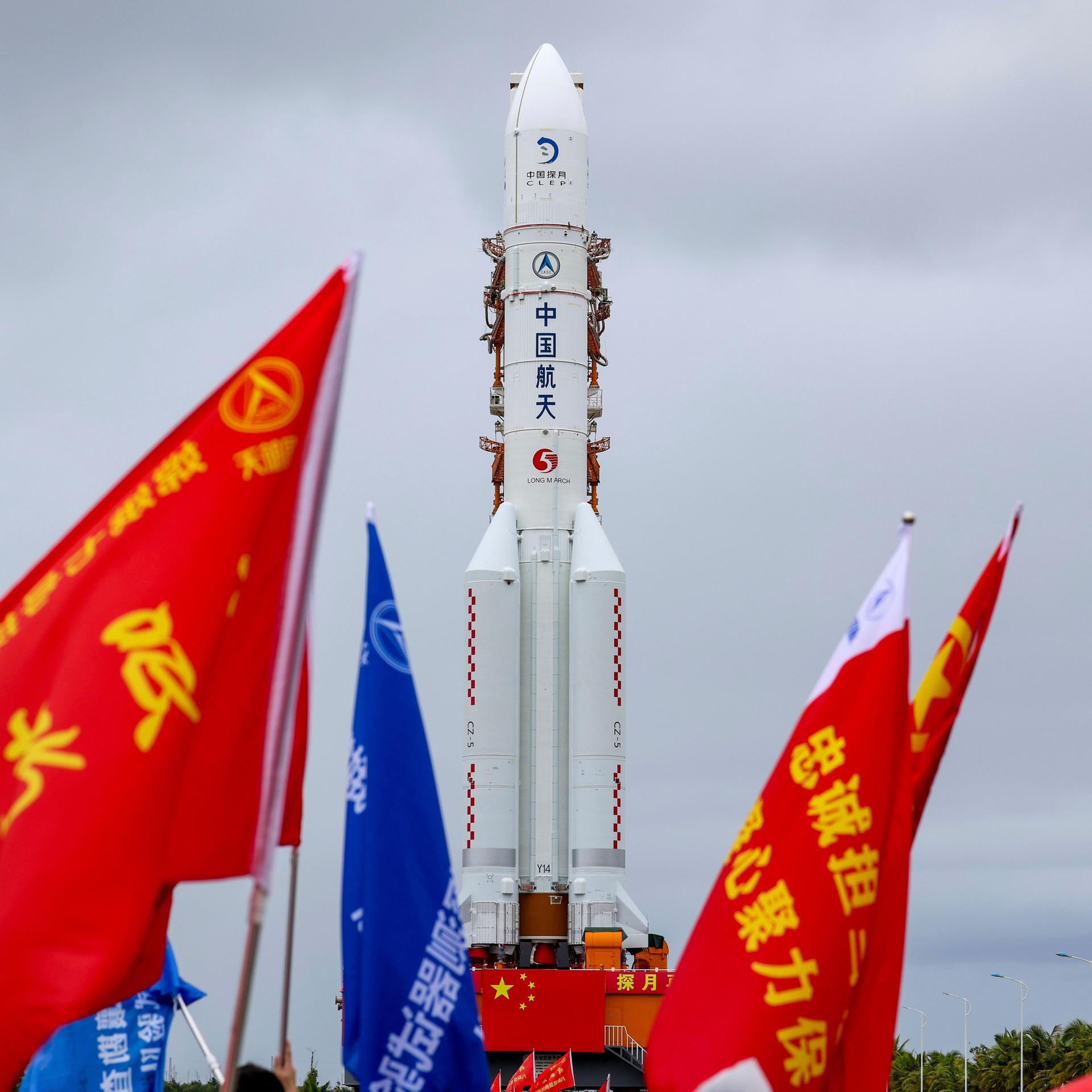 Die Mondsonde Chang'e-7 und ihre Trägerrakete Langer Marsch-5 Y14 am 19. August 2026 in der südchinesischen Provinz Hainan. Im Vordergrund sind rote und blaue Fahnen mit chinesischen Schriftzeichen.