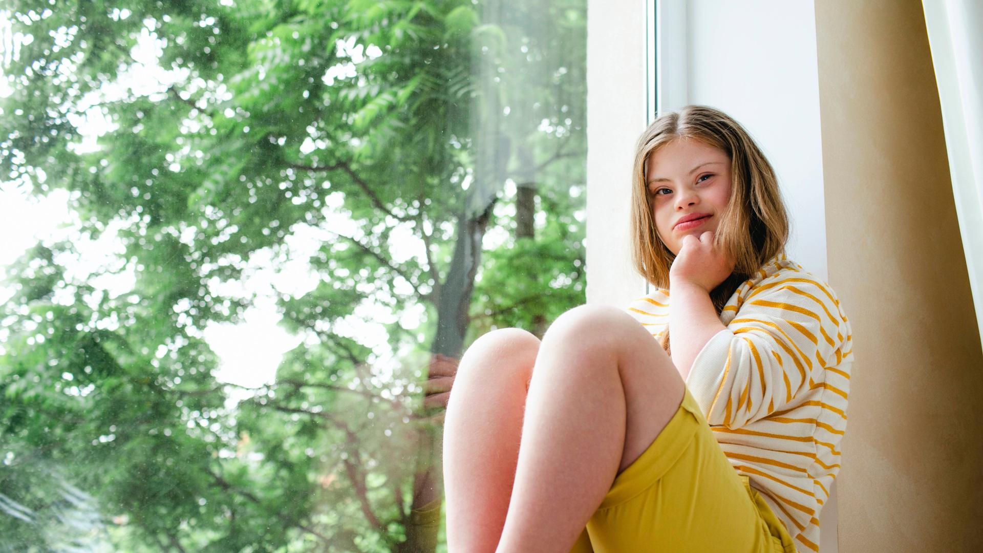 Ein Mädchen mit Down-Syndrom sitzt am Fenster, es trägt eine gelbe Shorts und einen Pullover mit gelben Streifen.
