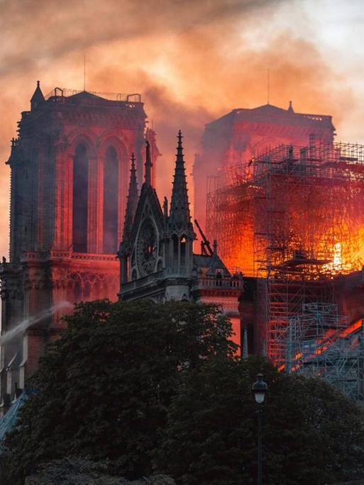 Eine Aufnahme aus dem Film "Notre-Dame in Flammen". Die bekannte Pariser Kirche brennt, die Flammen schlagen durch den Dachstuhl, am Rand wird versucht, mit Wasser zu löschen.