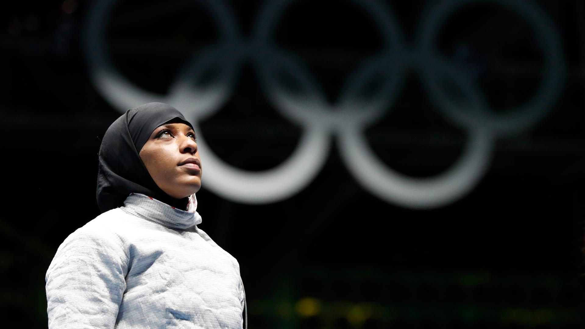 Eine Sportlerin mit einem Kopftuch steht vor den Olympischen Ringen.