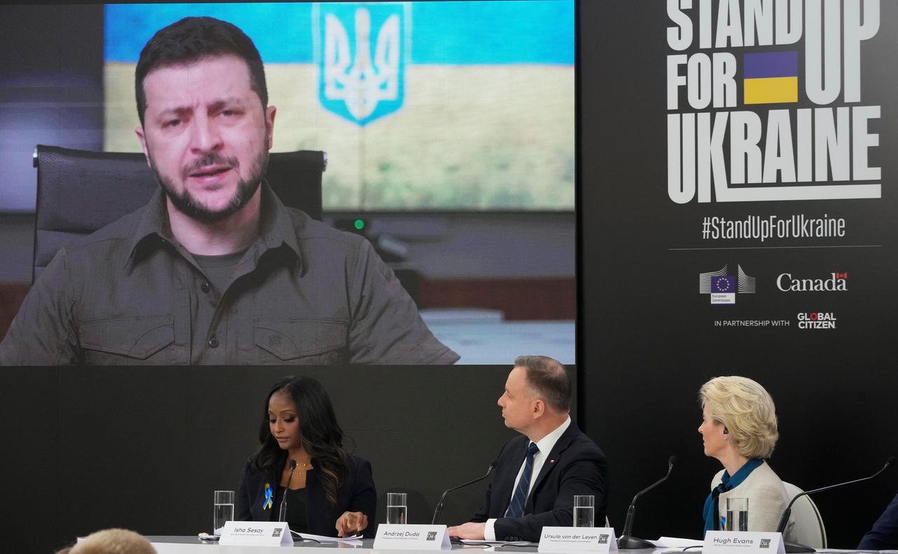 Zwei Frauen und ein Mann sitzen auf einem Podium und schauen auf eine Videoleinwand. Rechts ist ein großer Schriftzug zu sehen, der übersetzt "Steh' auf für die Ukraine" lautet.