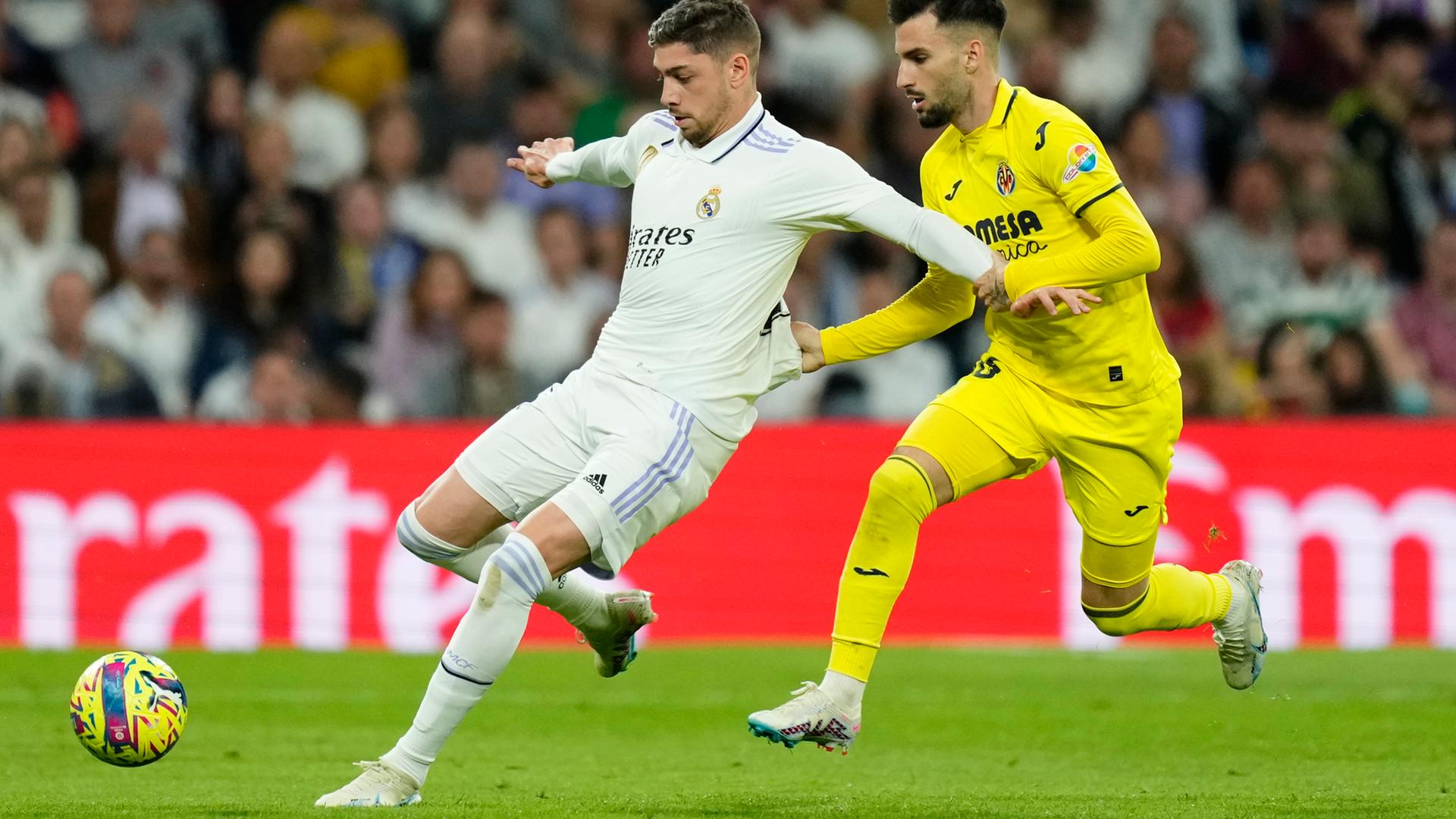 Zwei Fußballspieler kämpfen um den Ball. Der eine, Alex Baena von Villareal, zieht den anderen, Real Madrids Federico Valverde, am T-Shirt.