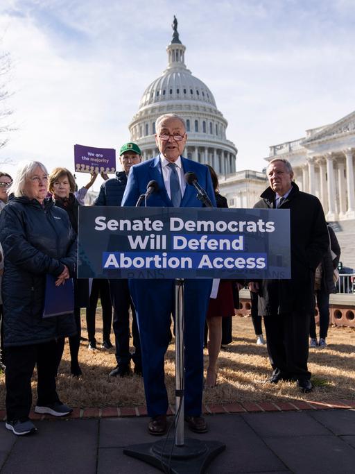 Der demokratische Senatsmehrheitsführer Chuck Schumer steht in einer Gruppe Menschen hinter einem Rednerpult vor dem US Kongress. Vor ihm steht ein Schild: "Senate Democrats will defend abortion access."
