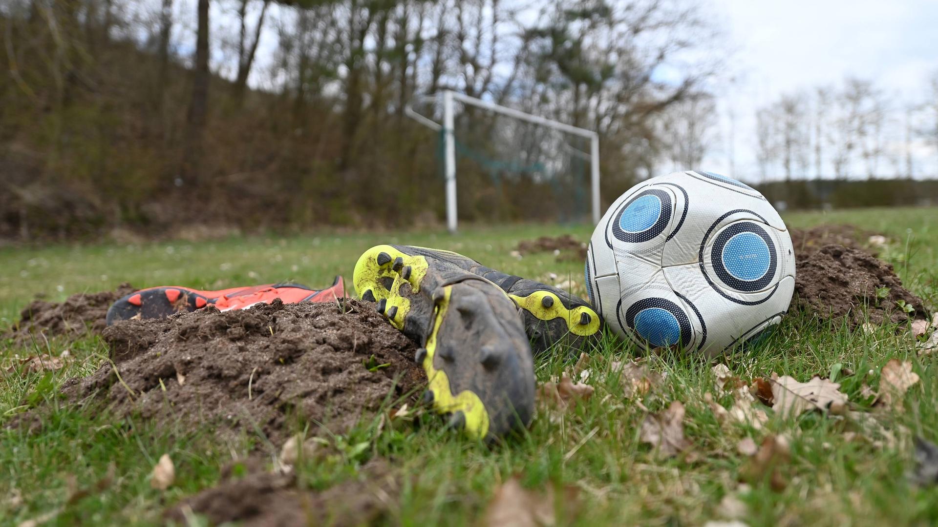 Symbolbild Fußball. Ein Spielball ist platt, ohne Luft. Fussballschuhe liegen auf einem schlechten Rasen mit Maulwurfshügeln