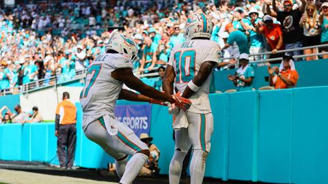 Jayleen Waddle hält seinem Mitspieler Tyreek Hill die Hände auf den Rücken, als würde er Hill Handschellen anlegen. Die beiden stehen in einem Football-Stadion, im Hintergrund feiern Fans den Punktgewinn.