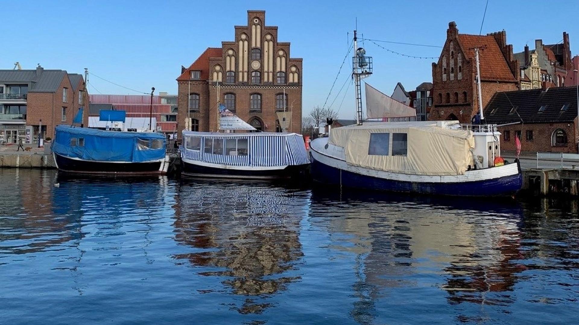 Blick auf Hafengelände in Wismar mit Booten im Vordergrund, die vor historischen Gebäuden ankern.