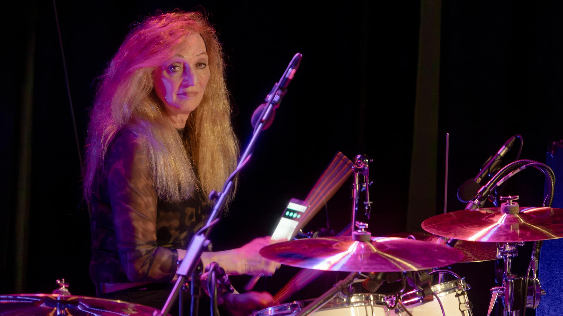 Schlagzeugerin Tina Powileit, Drummerin der Band Die Seilschaft, live in der Börse Coswig. Sie sitzt hinter ihrem Drumset und lächelt, das Licht im Hintergrund schimmert rot-blau. 