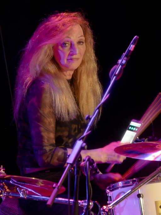 Schlagzeugerin Tina Powileit, Drummerin der Band Die Seilschaft, live in der Börse Coswig. Sie sitzt hinter ihrem Drumset und lächelt, das Licht im Hintergrund schimmert rot-blau. 