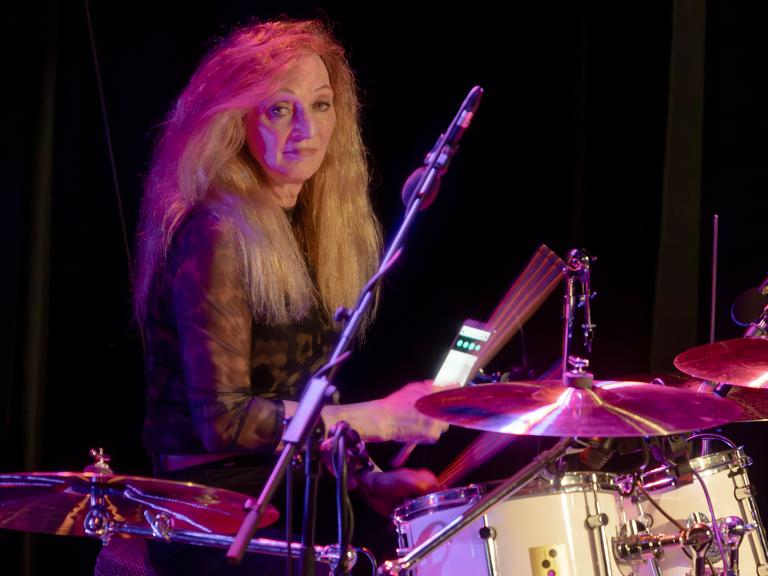 Schlagzeugerin Tina Powileit, Drummerin der Band Die Seilschaft, live in der Börse Coswig. Sie sitzt hinter ihrem Drumset und lächelt, das Licht im Hintergrund schimmert rot-blau. 